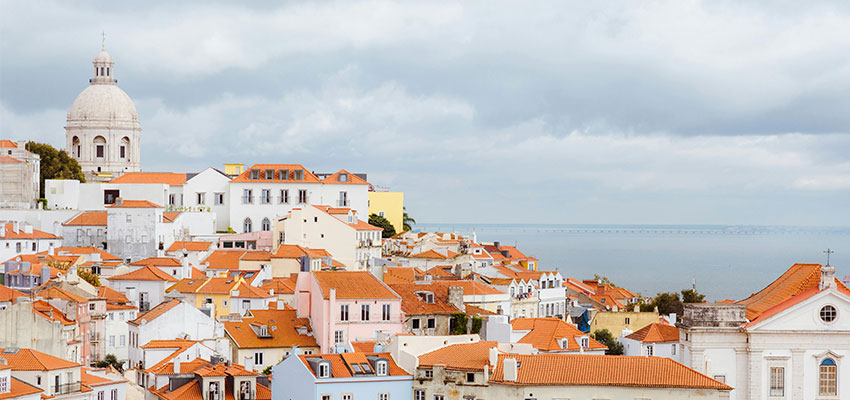 Vista panorâmica de Lisboa com casas de telhados alaranjados e o rio ao fundo em dia nublado