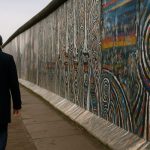 Homem caminhando sozinho ao longo do Muro de Berlim grafitado, simbolizando reflexão e aprendizado